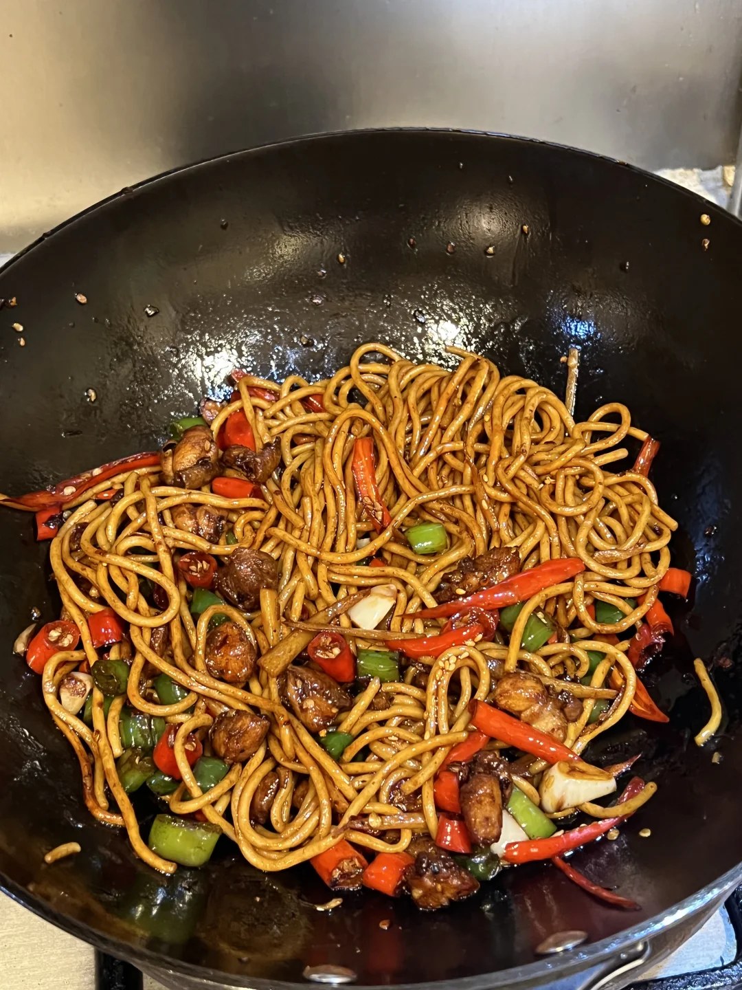 Brown noodles with chicken pieces and green and red chile slices in a pan.