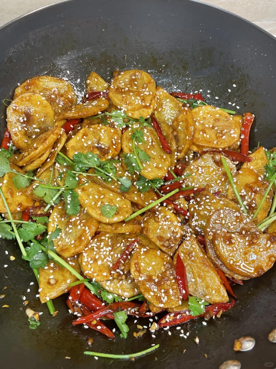 Golden potato slices covered with chile sauce, cilantro and sesame in a dark pan.