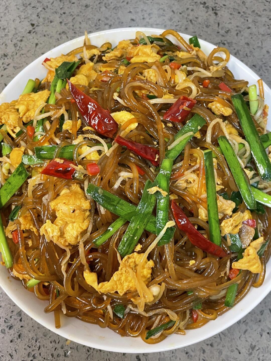 Brown glass-like noodles with green chives, bean sprouts and egg pieces on a white plate.