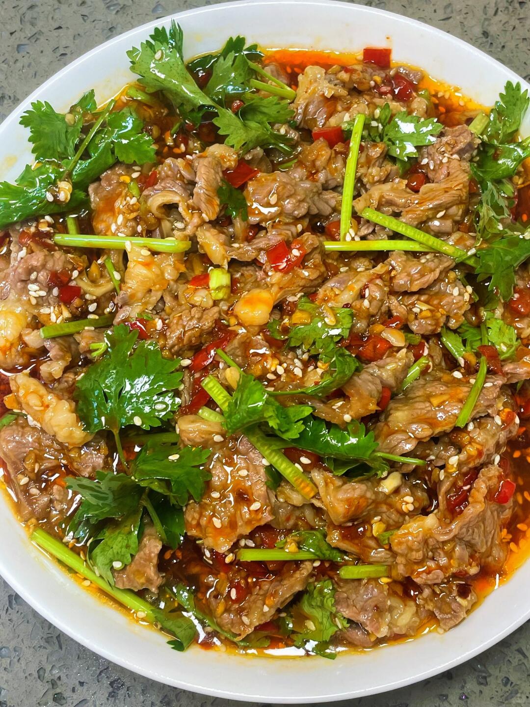 Beef slices covered with cilantro, sesame seeds and chopped red chiles in a shallow white bowl.