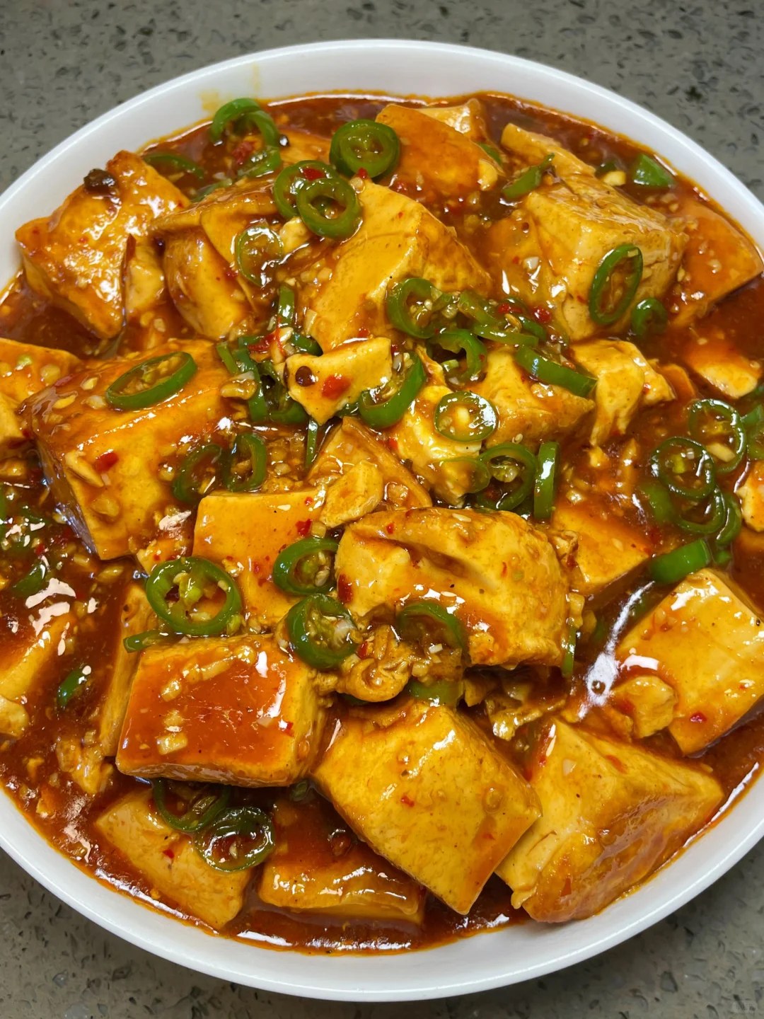 Golden tofu cubes in red sauce with chopped green chiles and scallions in a white bowl.