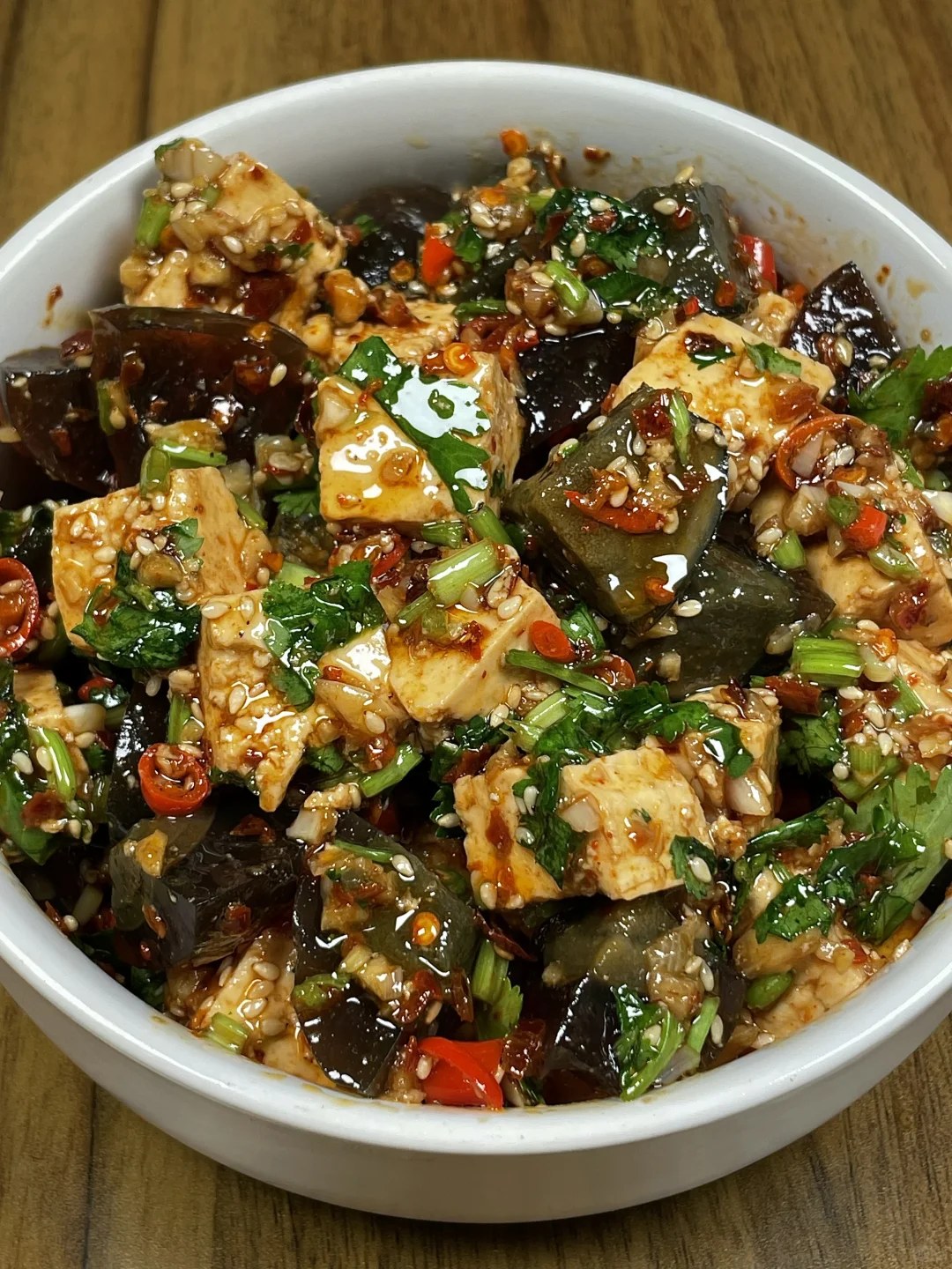 Tofu and dark century egg pieces topped with red chiles, green herbs and sesame in a white bowl.