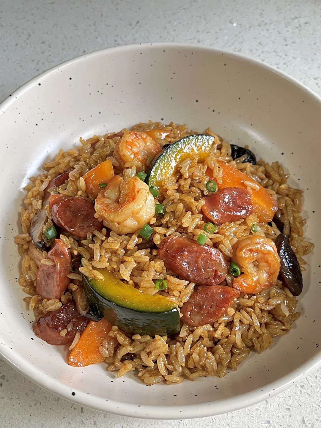 Brown seasoned rice topped with shrimp, orange pumpkin and Chinese sausage pieces in a white bowl.